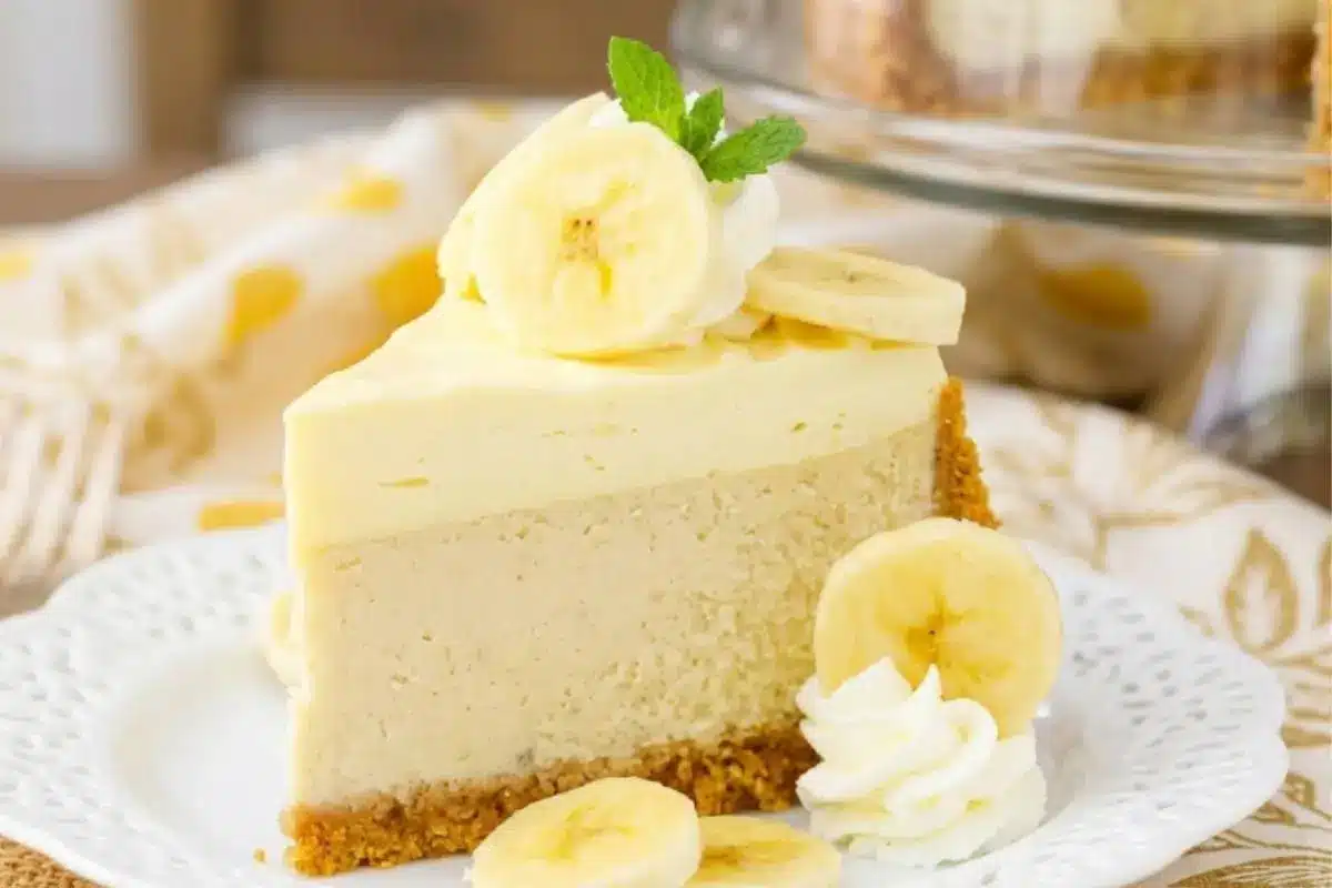 The Best Homemade Banana Cheesecake from Scratch 3 A comforting slice of creamy banana cheesecake featuring a thick, golden-brown cookie crust, topped with fresh banana slices and whipped cream on a rustic white plate.