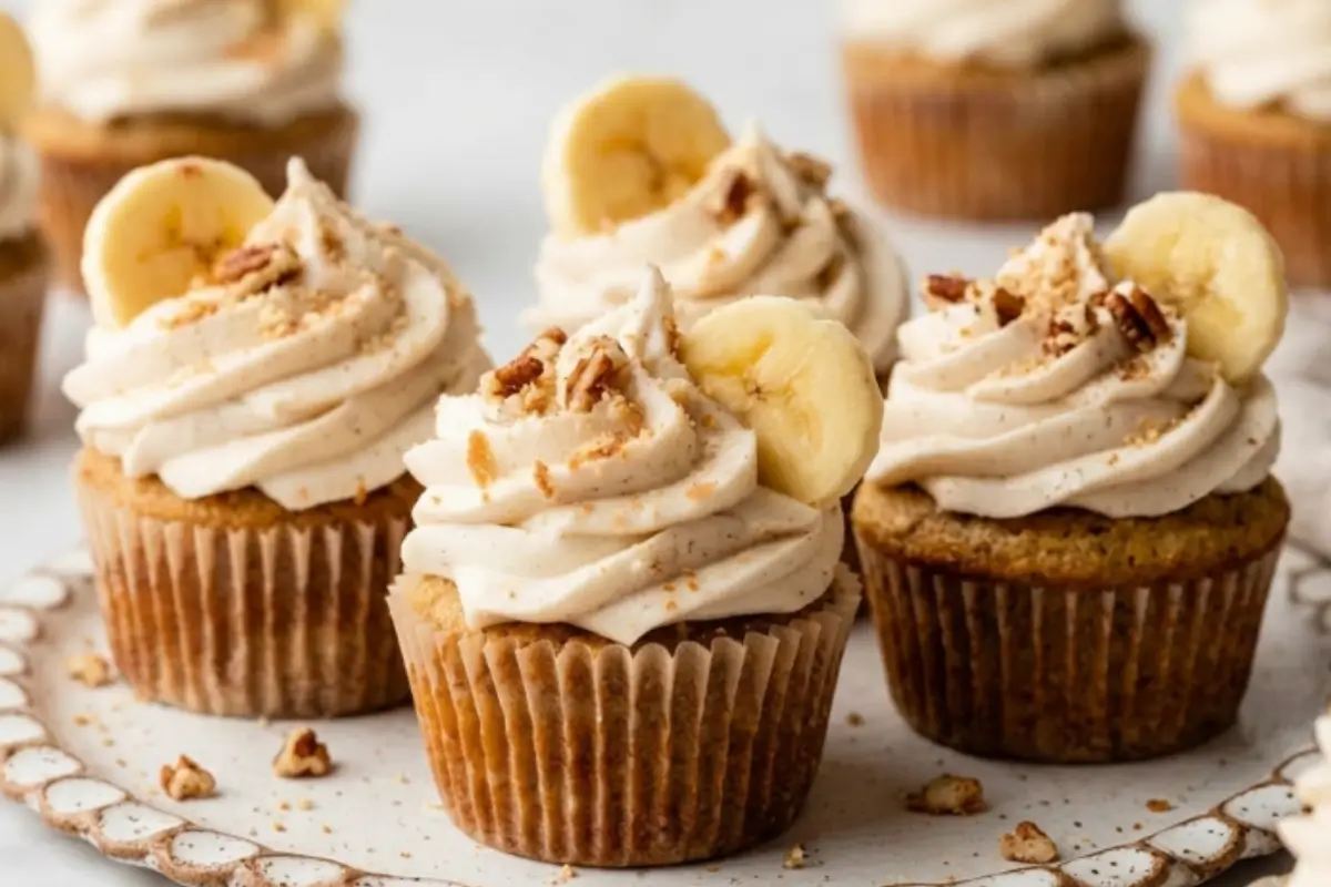 My Nonna's Ultimate Banana Cupcake Recipe: A Comforting Classic 3 A close up of four beautifully rustic banana cupcakes topped with creamy frosting, a fresh banana slice, and chopped pecans, sitting on a scalloped ceramic plate to showcase this easy banana cupcake recipe.
