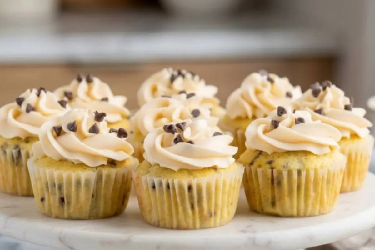 A beautiful batch of freshly baked chocolate chip cupcakes, generously swirled with creamy vanilla frosting and sprinkled with mini chocolate chips, sitting together on a round marble cake stand.