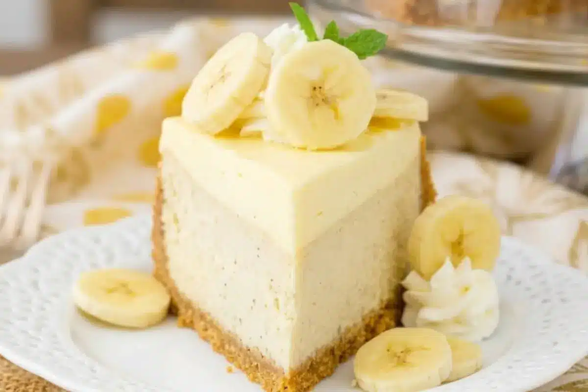Easy Banana Cheesecake_ A beautiful slice of easy banana cheesecake resting on a delicate white patterned plate, featuring a classic crumb crust, a rich, creamy banana center, and finished with fresh banana slices, sweet whipped cream, and a sprig of mint.