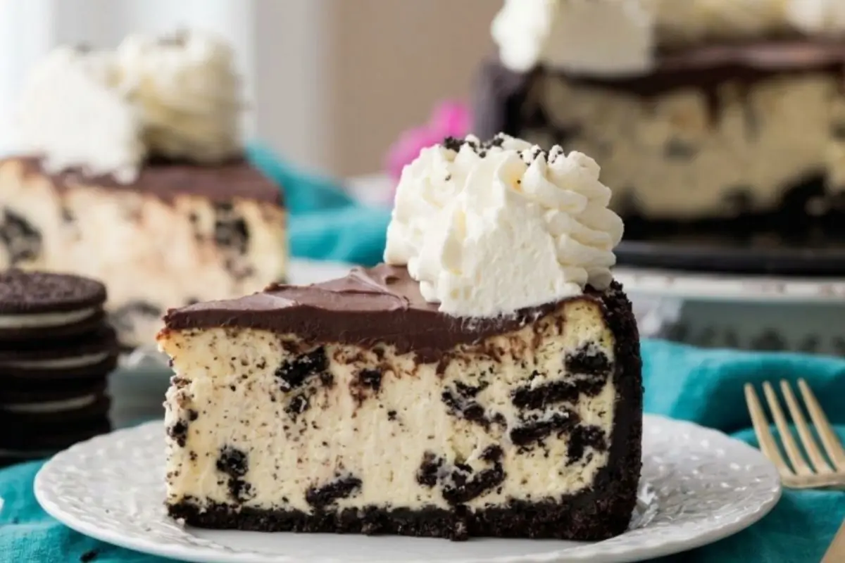 Easy Oreo Cheesecake A decadent slice of easy Oreo cheesecake resting on a white vintage plate with a gold fork, showing a thick chocolate cookie crust, a creamy center packed with crushed cookies, and a rich chocolate ganache topped with fresh whipped cream.