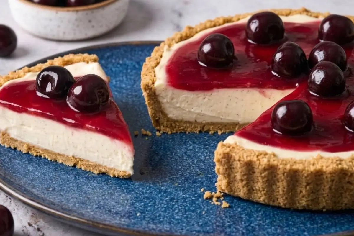 Easy Philadelphia No-Bake Cheesecake A beautiful slice of easy Philadelphia no-bake cheesecake resting on a rustic blue speckled plate alongside the remaining tart, featuring a golden crumb crust, a creamy vanilla-flecked center, and a vibrant cherry topping set against a light-grey textured concrete backdrop.