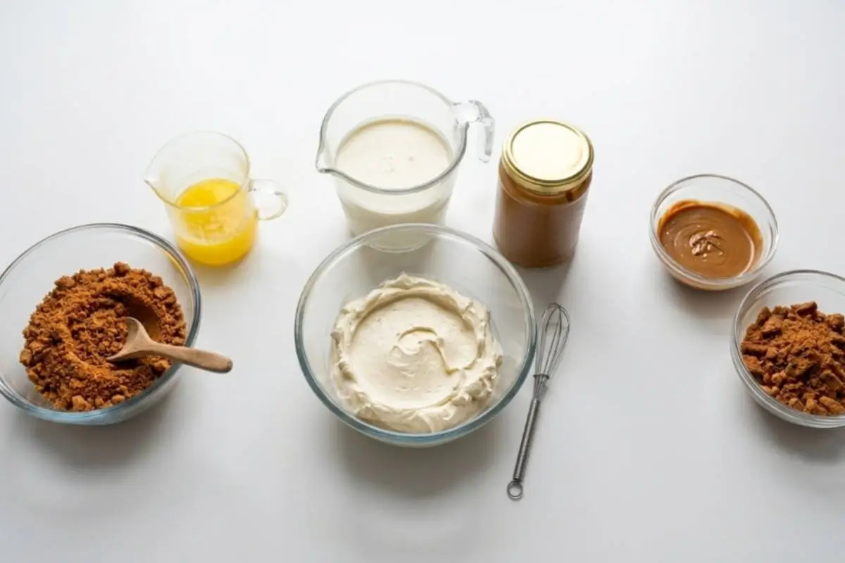 Ingredients For Biscoff Cheesecake An overhead view showing the gathered ingredients for biscoff cheesecake on a clean white surface, featuring crushed Biscoff cookies in a glass bowl with a rustic wooden spoon, a pitcher of heavy cream, a bowl of smooth cream cheese, melted butter, and a jar of rich cookie butter next to a wire whisk.