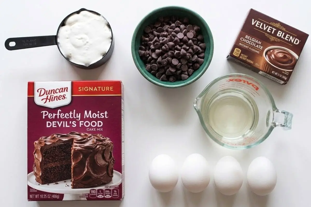 _Ingredients For Chocolate Chip Bundt Cake A top-down view of the simple, accessible ingredients needed to bake a comforting chocolate chip bundt cake, including a box of Devil's food cake mix, instant chocolate pudding, a bowl of chocolate chips, sour cream, water, and four fresh eggs arranged neatly on a white surface.