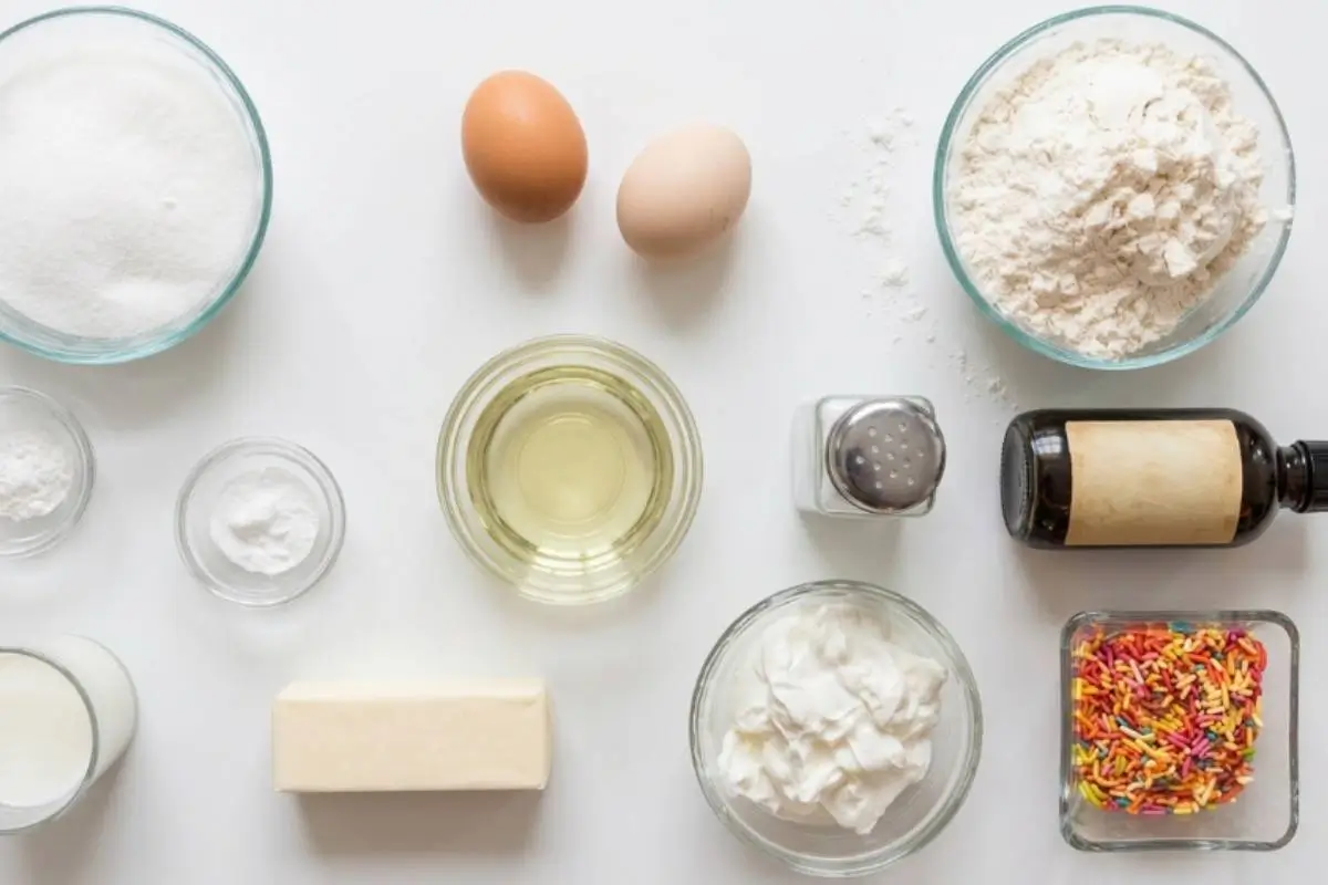 Key Ingredients For Birthday Cupcakes An overhead view of the simple, everyday ingredients needed to bake homemade birthday cupcakes, including bowls of flour, sugar, fresh eggs, butter, and vibrant rainbow sprinkles.