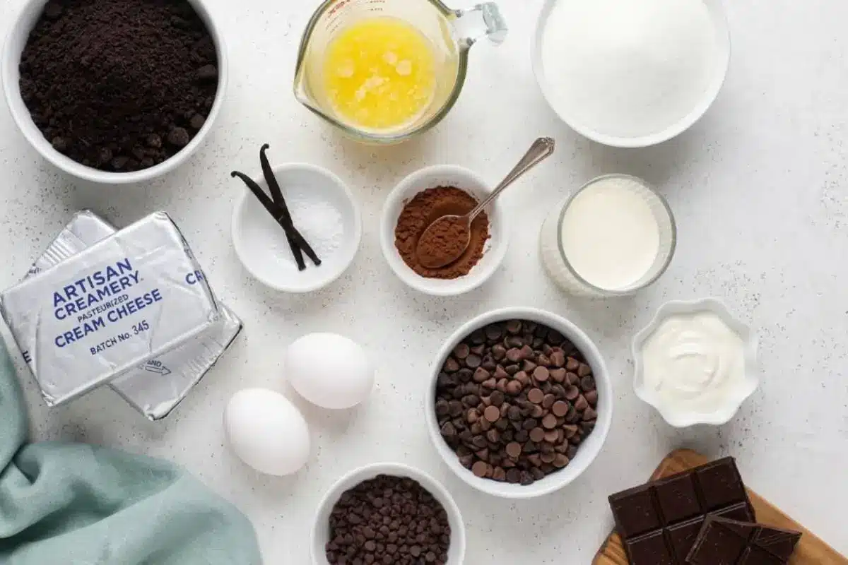 Key Ingredients For Chocolate Chip Cheesecake An overhead static angle of chocolate chip cheesecake ingredients, including artisan cream cheese, fresh eggs, chocolate chips, and vanilla, arranged on a light-grey textured concrete backdrop.