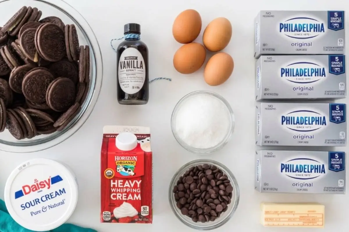 Key Ingredients For Oreo Cheesecake An overhead view of the simple, wholesome ingredients needed for an Oreo cheesecake, including a bowl of classic Oreo cookies, original cream cheese, fresh eggs, sour cream, and chocolate chips, resting on a bright kitchen counter.