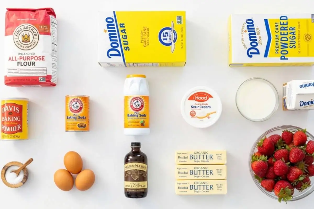 An inviting overhead view of the essential baking ingredients needed for a homemade strawberry cupcake recipe, beautifully arranged on a clean white surface and including a bowl of fresh red strawberries, flour, sugar, butter, eggs, milk, and vanilla extract.