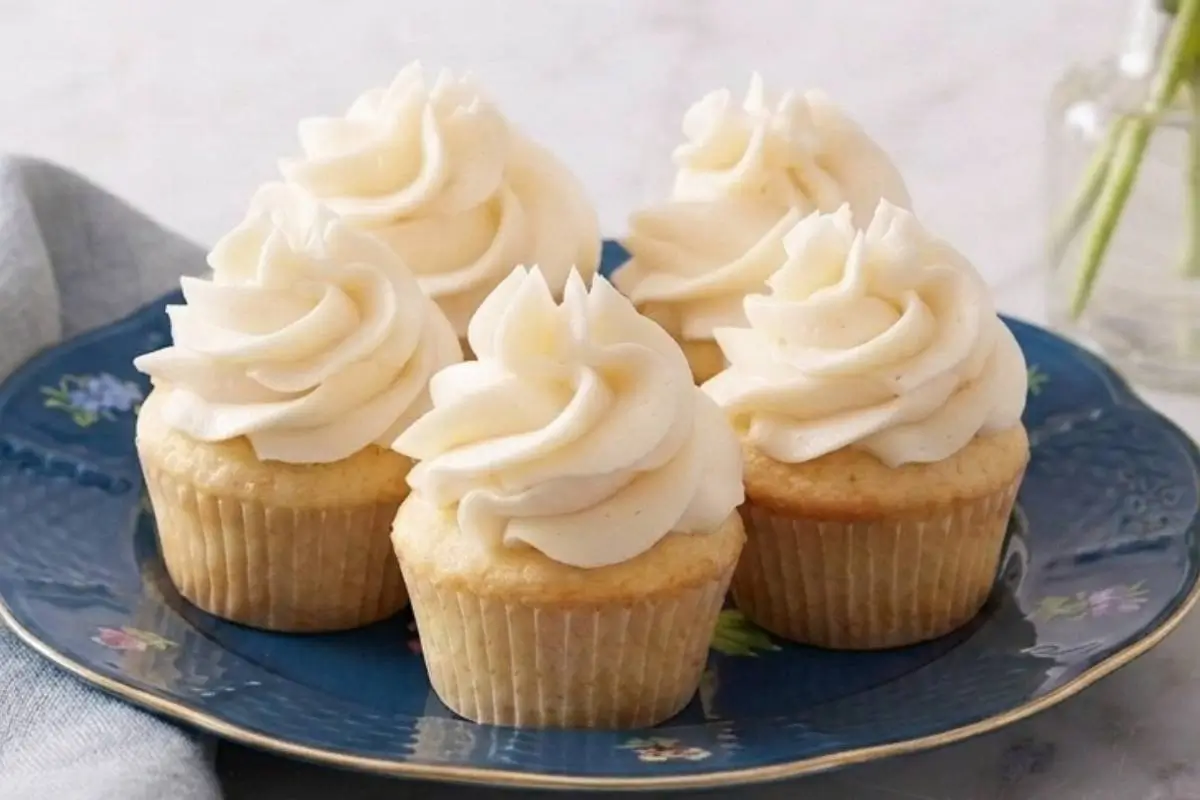 School-Safe Mini Cupcakes: An Easy, Nut-Free Recipe Kids Will Love 3 A cluster of five freshly baked mini cupcakes with generous swirls of creamy vanilla frosting, arranged on an elegant, dark blue floral-patterned plate.