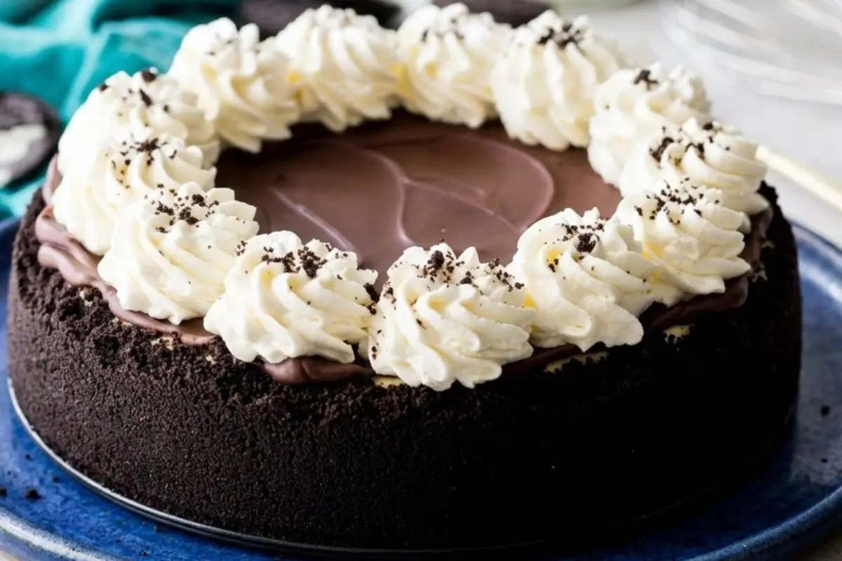 Easy Oreo Cheesecake with Chocolate Crust (No Bake/Baked) 3 A decadent Oreo cheesecake with a dark chocolate cookie crust, topped with a smooth chocolate ganache and beautiful swirls of whipped cream sprinkled with cookie crumbs, resting on a blue ceramic plate.