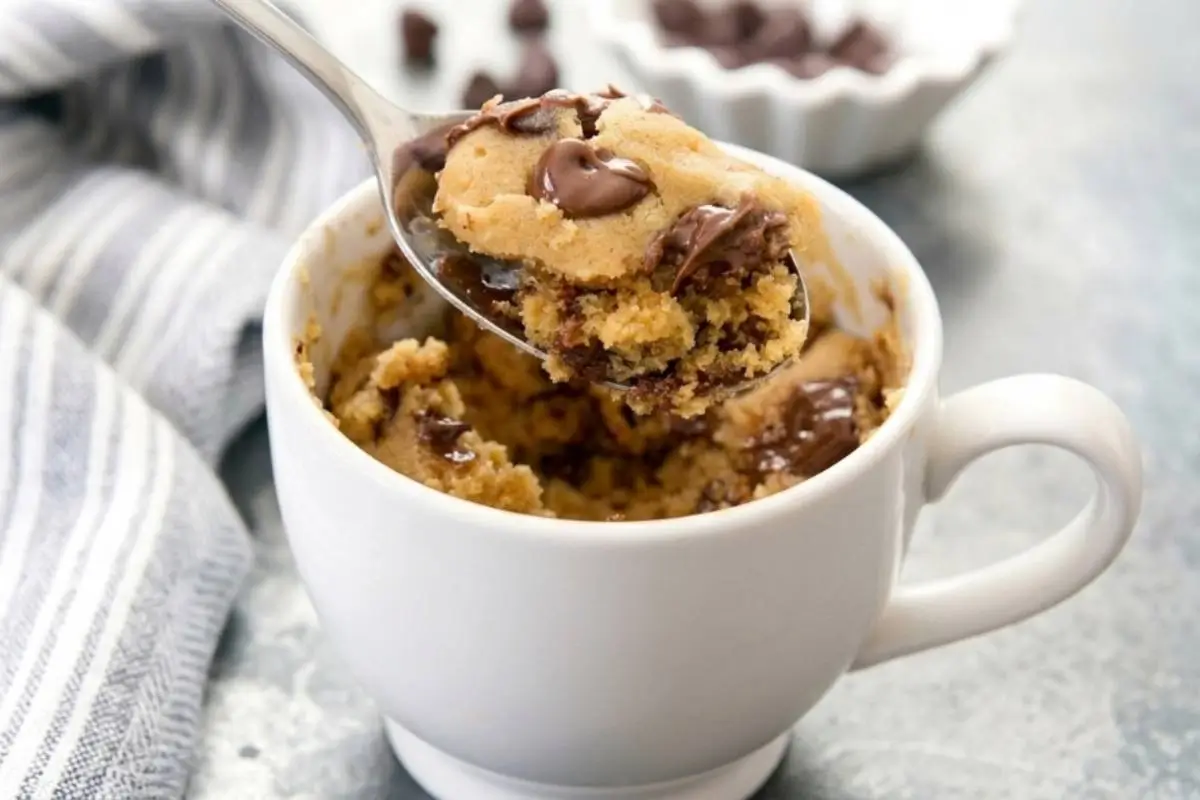 A silver spoon lifts a warm, gooey bite of baked chocolate chip mug cake from a cozy white ceramic mug, set against a rustic surface with a folded striped linen napkin and a small bowl of chocolate morsels in the background.