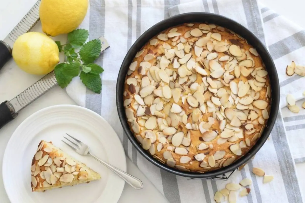 Storage Tips, Freezing For Your Almond Ricotta Cake A golden, freshly baked almond ricotta cake recipe completely covered in toasted sliced almonds inside a round baking pan, resting on a rustic striped linen towel next to bright yellow lemons, a zester, and a single cut slice on a white ceramic plate.