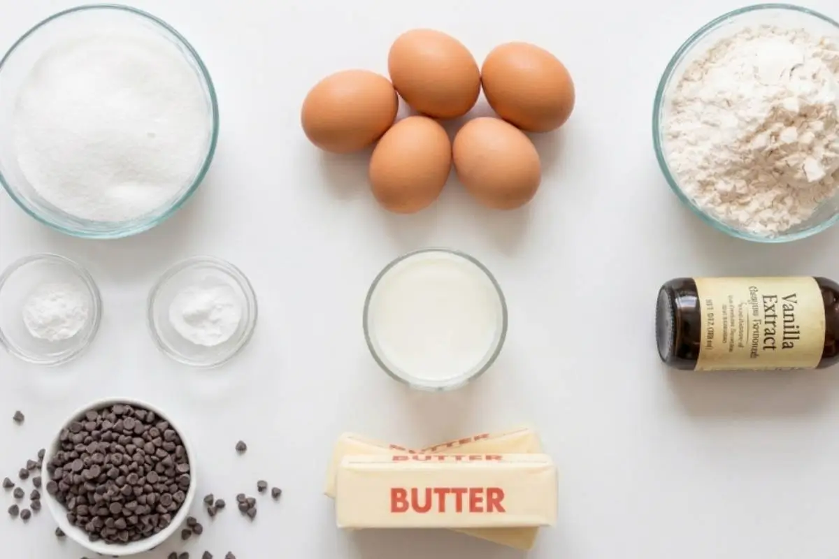 An inviting overhead view of the simple, everyday pantry ingredients needed to bake homemade chocolate chip cupcakes, beautifully arranged on a white surface and including flour, sugar, fresh brown eggs, butter, milk, vanilla extract, and a bowl of mini chocolate chips.