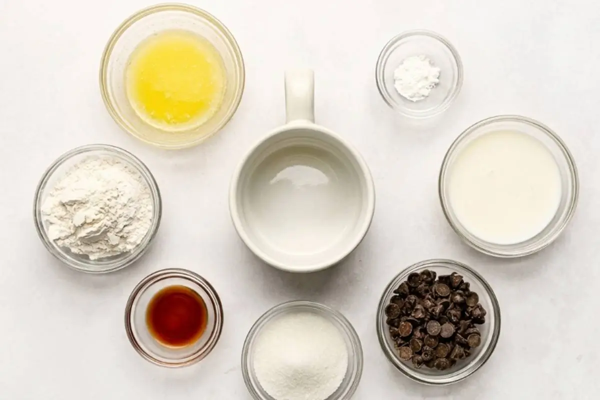A top-down view of the simple ingredients needed for a chocolate chip mug cake, including flour, sugar, milk, vanilla, and chocolate chips, naturally arranged in small glass bowls around a cozy white ceramic mug on a light textured surface.