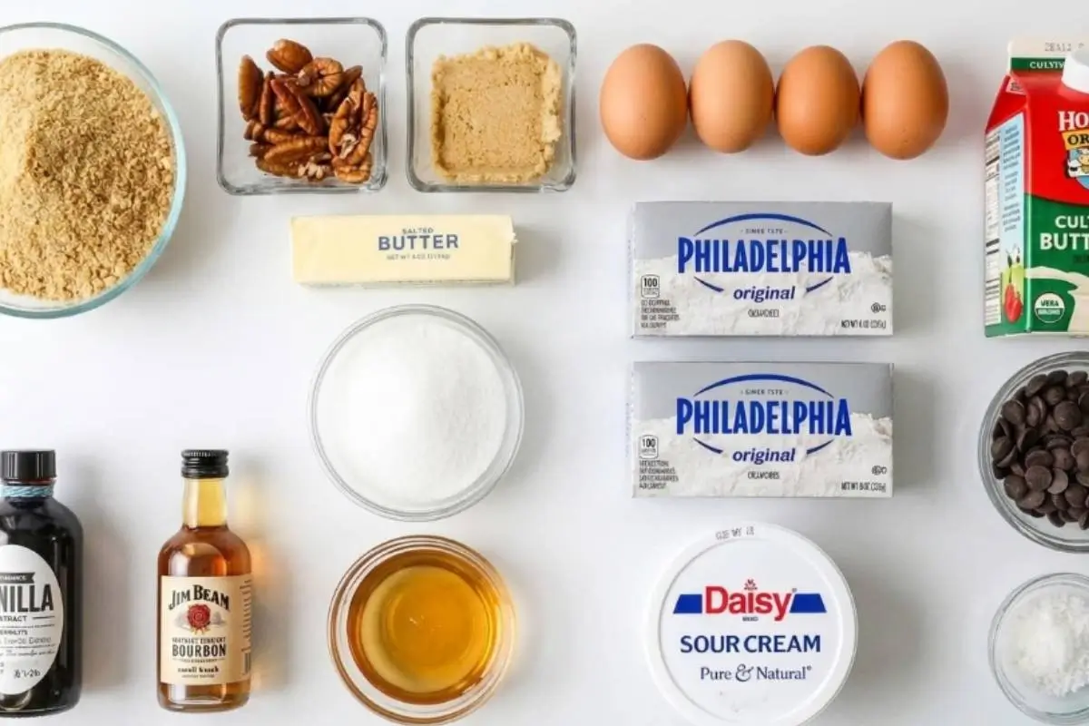 The Key Ingredients For The Decadent Turtle Cheesecake A beautifully organized overhead flat lay of the essential ingredients for a decadent turtle cheesecake laid out on a white counter, featuring blocks of cream cheese, fresh pecans, brown sugar, eggs, butter, and chocolate chips.