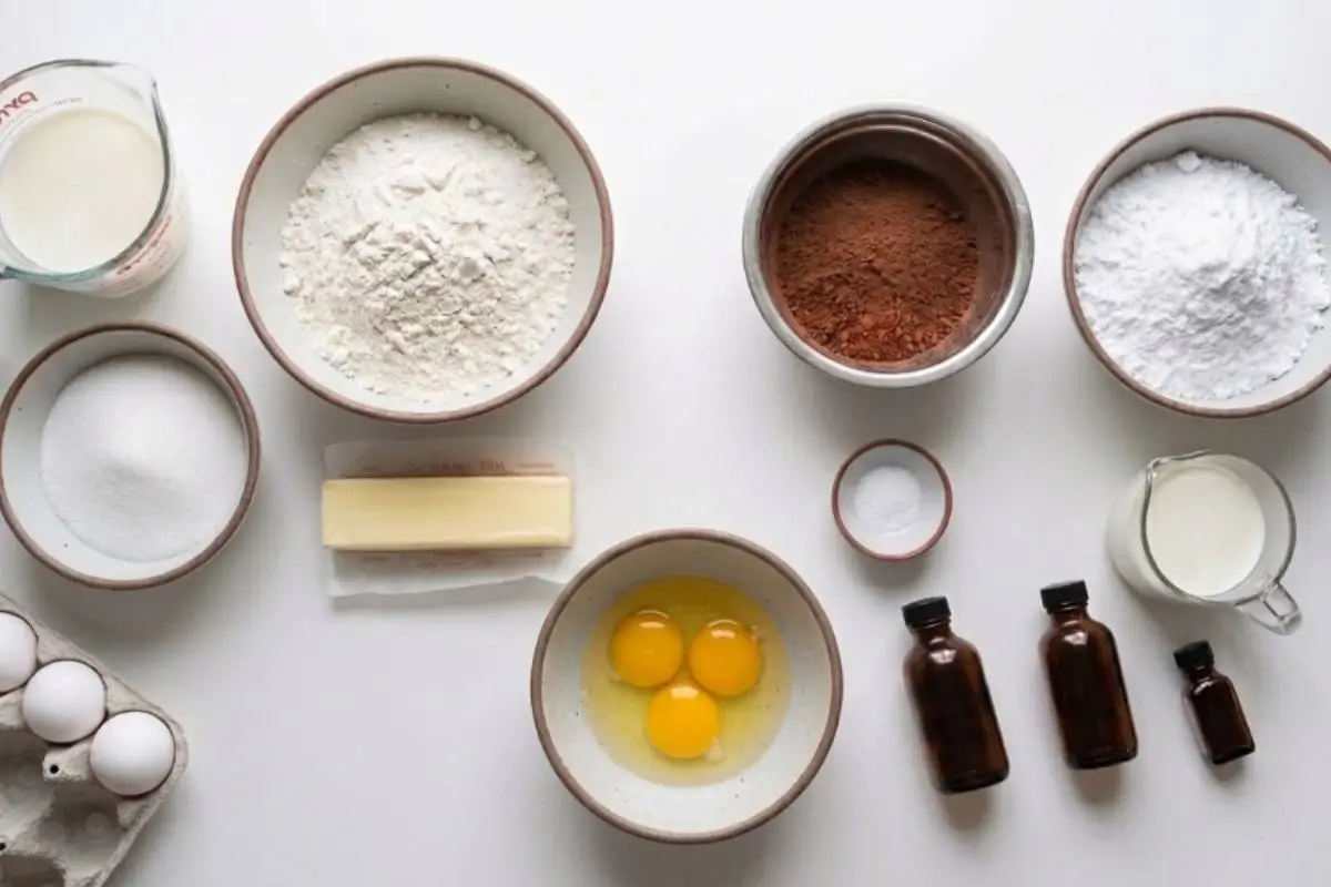 The Key Ingredients For These Gluten-Free Cupcakes A top-down view of simple baking ingredients for gluten-free cupcakes, including fresh eggs, butter, cocoa powder, and flour, arranged beautifully in warm, rustic ceramic bowls on a white surface.