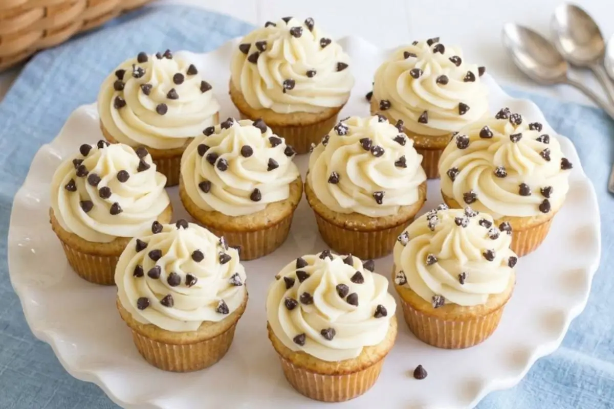 How to Bake Foolproof Cannoli Cupcakes (Just Like Nonna's) 3 A beautiful batch of freshly baked cannoli cupcakes on a white scalloped cake stand, generously piped with sweet ricotta frosting and sprinkled with mini chocolate chips, resting gently on a soft blue linen cloth.