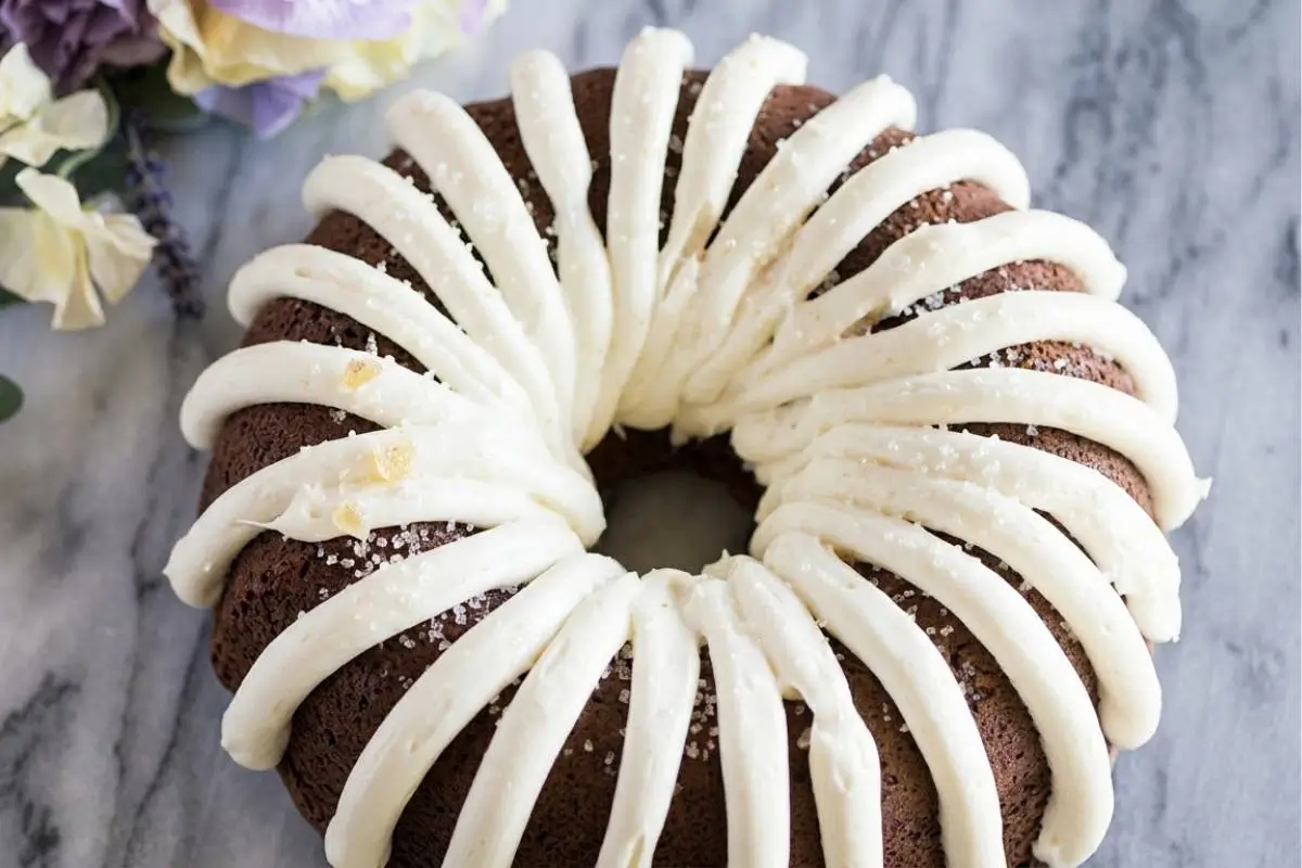 Simply the Best Chocolate Chip Bundt Cake 3 A rich, dark chocolate chip bundt cake elegantly decorated with thick ribbons of white frosting and sparkling coarse sugar, resting on a marble surface beside soft purple and yellow flowers.