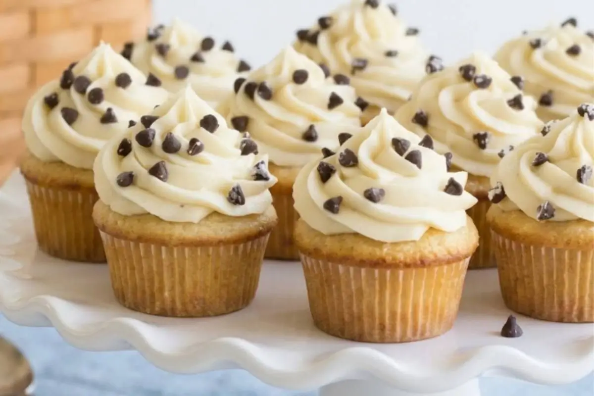 A beautiful batch of soft, rustic baked cannoli cookie cups, each generously crowned with a swirl of sweet ricotta frosting and dotted with mini chocolate chips, beautifully arranged on a white scalloped serving stand.