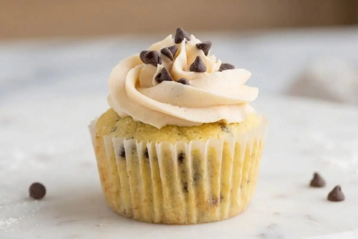 A soft, rustic chocolate chip cookie cup baked in a simple paper liner, crowned with a generous swirl of sweet frosting and dotted with mini chocolate chips, sitting on a softly lit, cozy marble surface.
