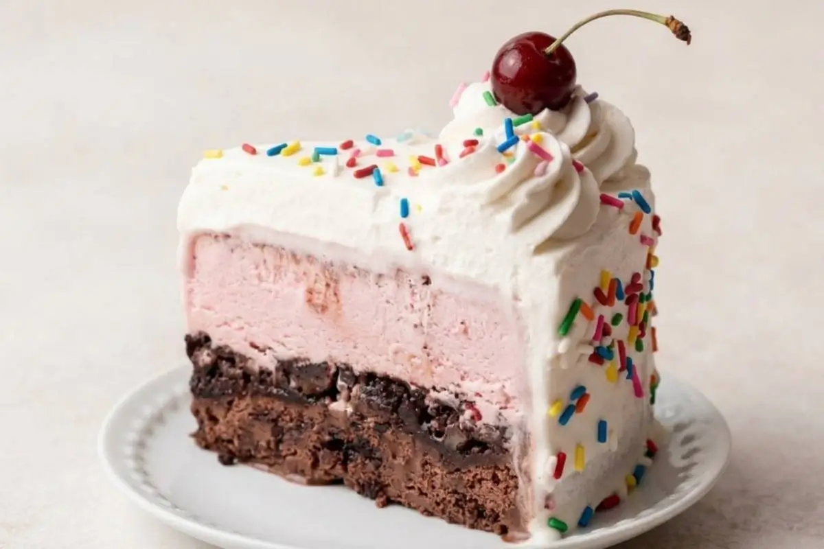 foolproof Ice Cream Cake A thick, colorful slice of a layered ice cream cookie stack, featuring a fudgy chocolate cookie base, a thick layer of strawberry ice cream, and fluffy whipped cream topped with rainbow sprinkles and a fresh cherry, resting on a simple white plate.