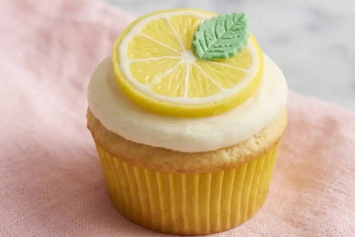 A soft, bright baked lemon cookie cup resting in a simple paper liner, crowned with smooth white frosting, a decorative candy lemon slice, and a small green leaf, all sitting on a soft, warm pink linen napkin.