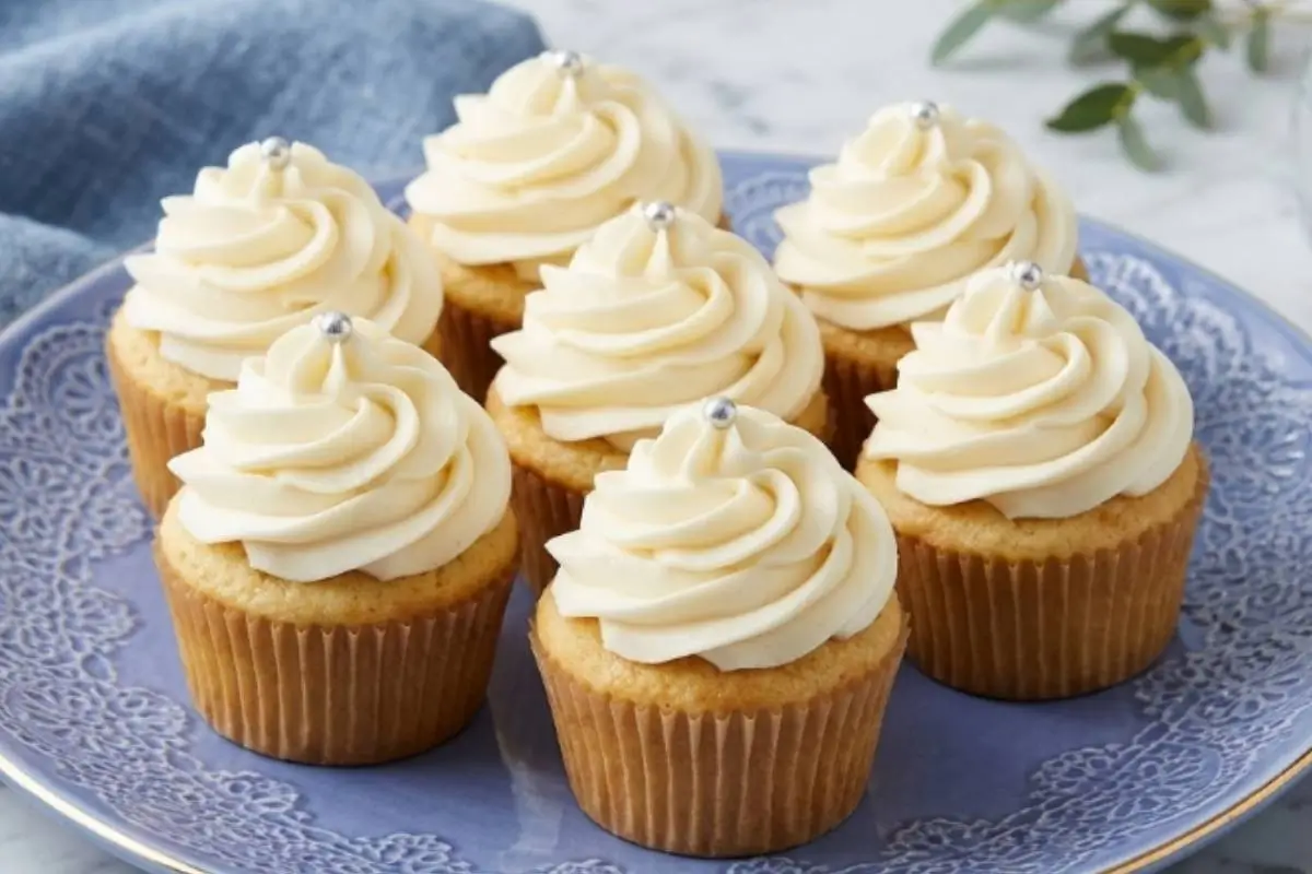 A beautiful batch of soft, baked vanilla mini cookie cups, each crowned with a generous swirl of sweet white frosting and a single silver sprinkle, arranged elegantly on a vintage-style blue ceramic plate.