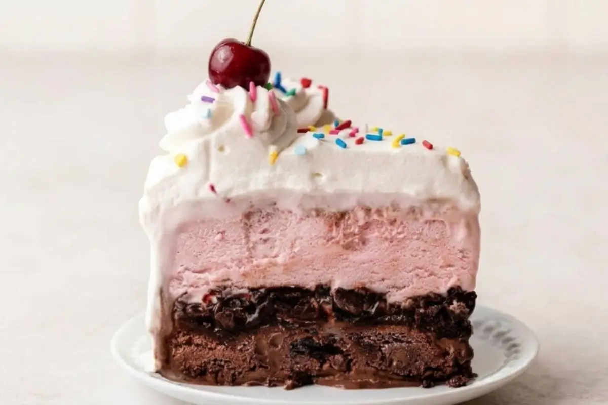The Best Traditional Homemade Ice Cream Cake Recipe 3 A beautiful, thick slice of our homemade ice cream cake recipe resting on a rustic white plate, showcasing nostalgic layers of rich chocolate ice cream, gooey fudge with crushed cookies, sweet pink strawberry ice cream, and fluffy whipped cream finished with colorful sprinkles and a fresh red cherry.