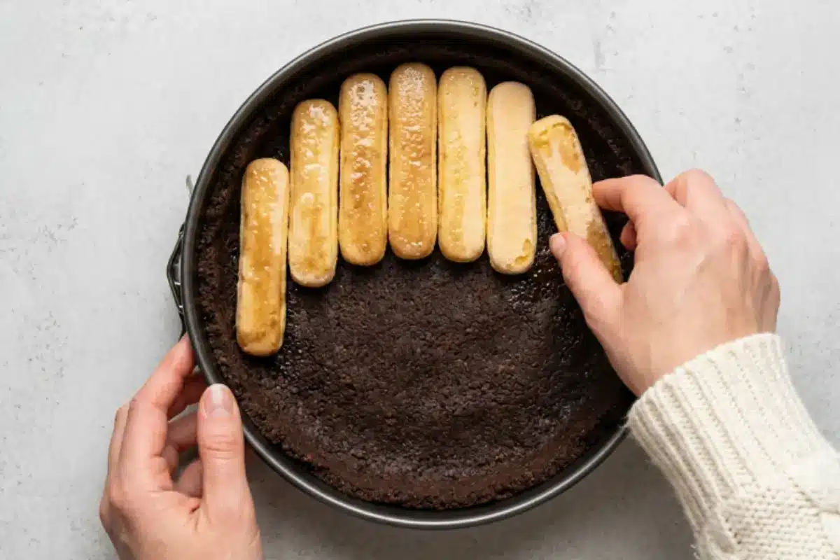 Build the filling base For Nutella Tiramisu Cake A warm, top-down view of a hand gently placing espresso-soaked ladyfingers onto a dark chocolate crumb crust inside a metal springform pan to build the base of a Nutella Tiramisu Cake.