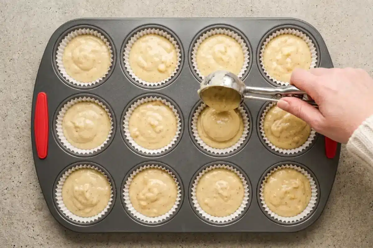 Elena’s No-Fail Tips For Your Banana Cupcakes A top-down view showing a hand using a metal scoop to gently drop batter into a lined muffin tin, a reassuring and easy step for baking our classic banana cupcakes recipe.