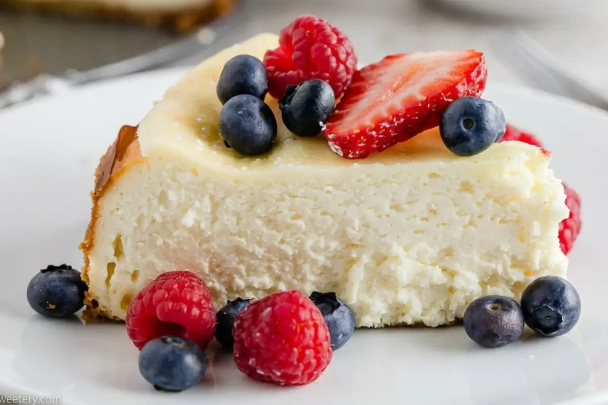 Gluten-Free Cheesecake 3 A close-up of a thick, creamy slice of gluten-free cheesecake topped with fresh raspberries, blueberries, and a sliced strawberry, served on a simple white plate.