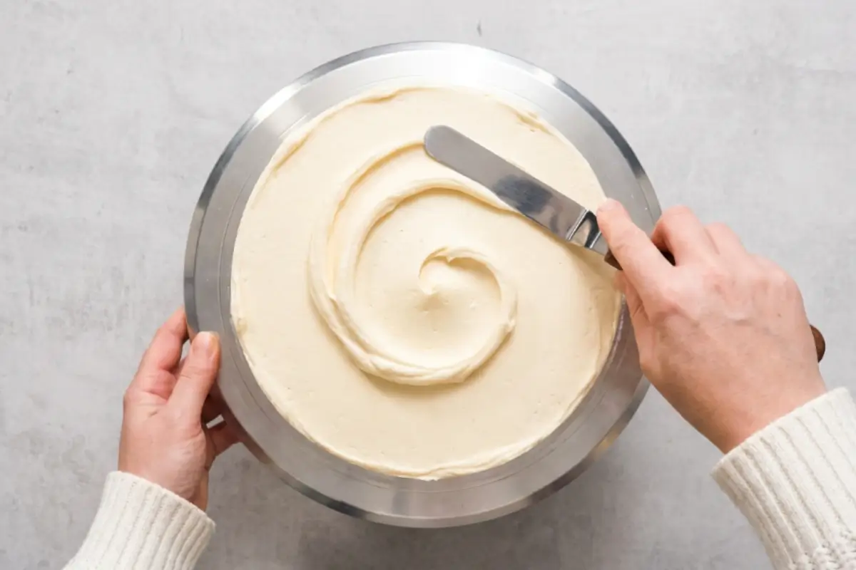 How to Smooth Frosting: Elena's No-Fail Method 2 A top-down view of hands in a cozy white sweater using an offset spatula to create a simple, rustic swirl in buttercream on a round cake, demonstrating an easy, forgiving way of how to smooth frosting.