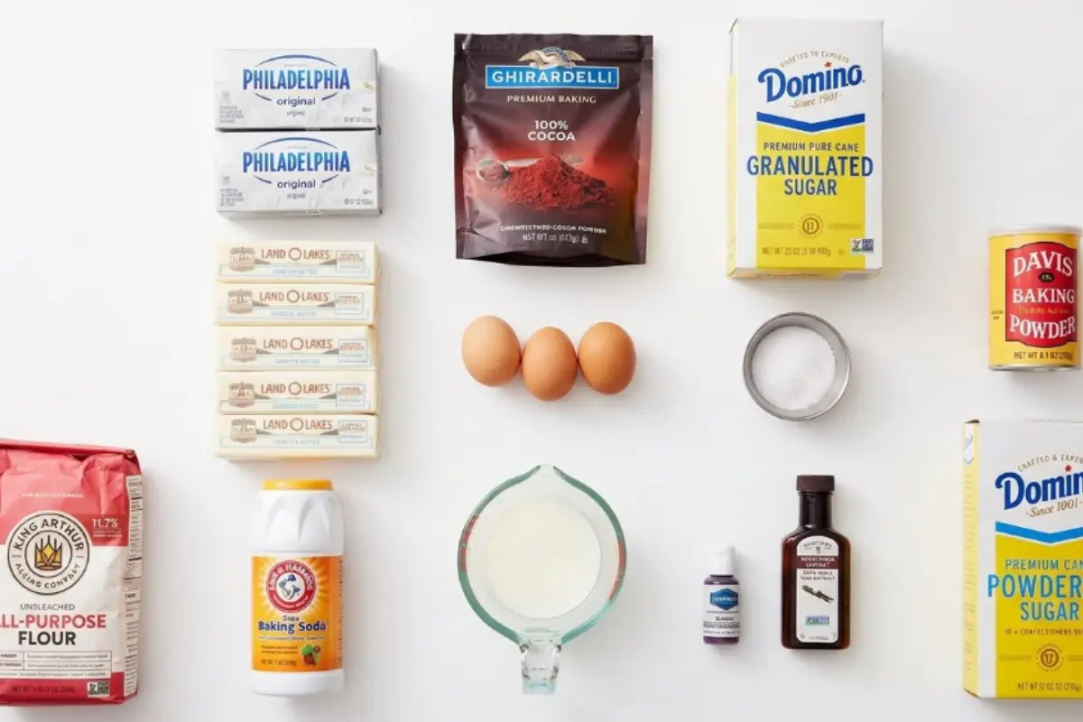 Key Ingredients For Red Velvet Cupcakes An overhead view of the classic baking ingredients needed to make red velvet cupcakes, including flour, cocoa powder, butter, eggs, buttermilk, sugar, and cream cheese, arranged neatly on a bright white surface.