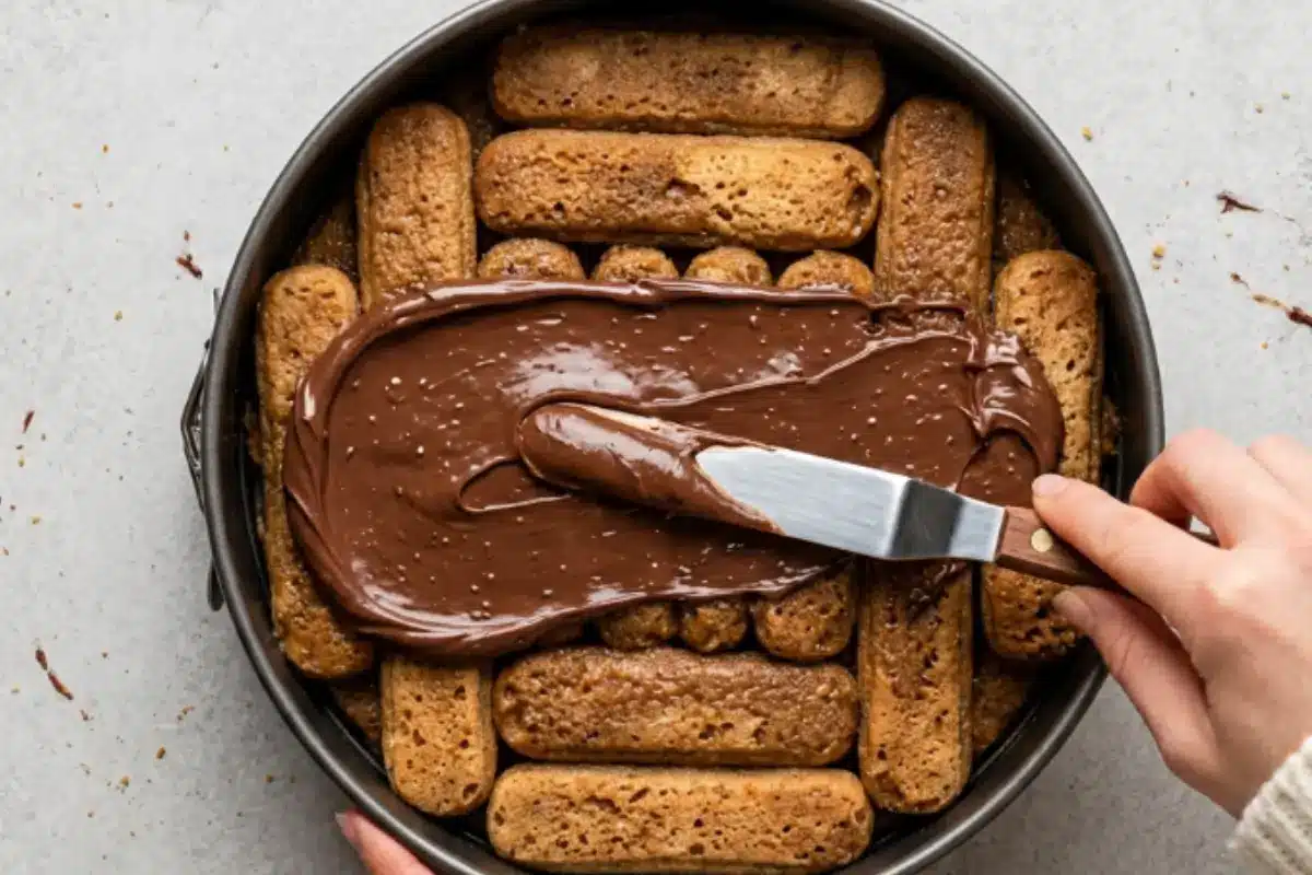 _No-Fail Tips For Nutella Tiramisu Cake A top-down view of a hand using a rustic, wooden-handled offset spatula to gently spread a thick, glossy layer of chocolate hazelnut spread over coffee-soaked ladyfingers inside a springform pan, showing the assembly of a Nutella Tiramisu Cake.