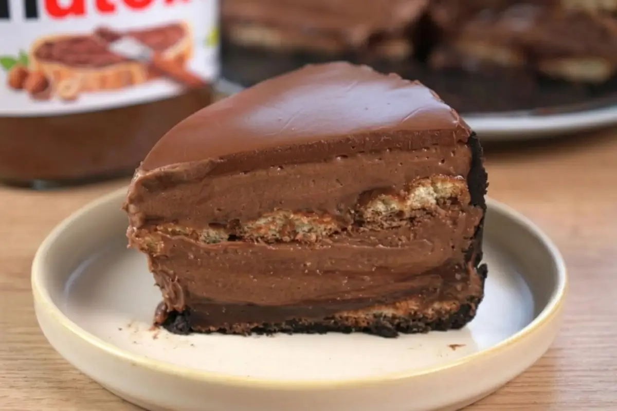 Nutella A beautiful, decadent slice of Nutella tiramisu cake resting on a simple, warm ceramic plate, showcasing its rustic dark chocolate crumb crust, espresso-soaked ladyfingers, and thick layers of creamy chocolate hazelnut mascarpone, with a jar of Nutella softly blurred in the background.