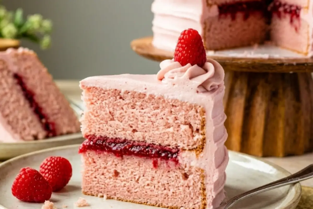 _Raspberry Cake recipe A thick, beautiful slice of a two-layer homemade raspberry cake recipe featuring a glossy, dark red berry jam filling and fluffy pink buttercream, served on a rustic ceramic plate with a silver dessert fork.