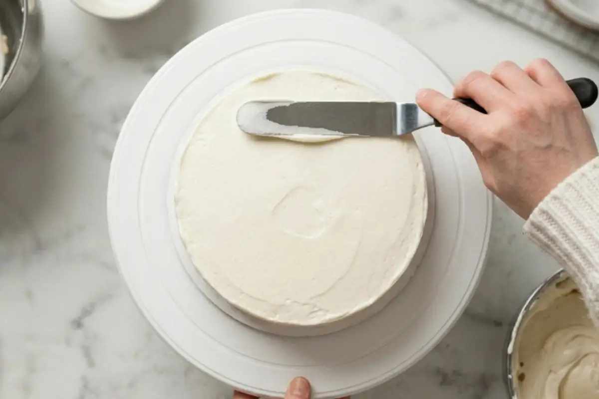 The Final Frosting A baker wearing a cozy cream sweater uses an offset spatula to gracefully smooth a thick, creamy final layer of white icing to beautifully frost a cake.