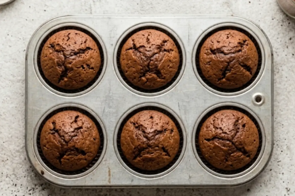 Tips for Chocolate Raspberry Cupcakes An overhead view of six freshly baked chocolate raspberry cupcakes resting in a well-loved metal muffin tin on a rustic kitchen counter, showcasing their perfectly cracked, domed tops.