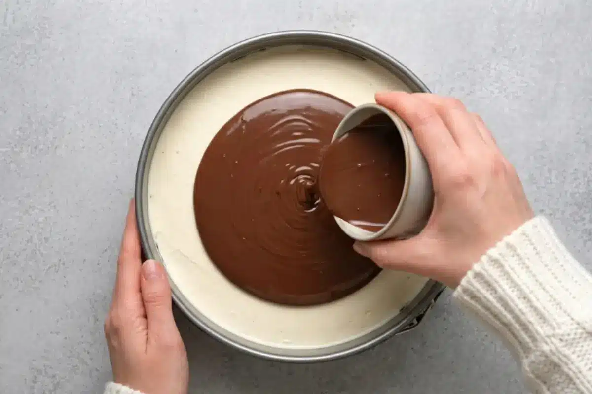 Why This Nutella A top-down view of a warm, cozy kitchen scene where a hand is pouring a glossy, rich pool of chocolate hazelnut spread from a small rustic bowl over a smooth white mascarpone layer in a springform pan for a Nutella Tiramisu Cake.