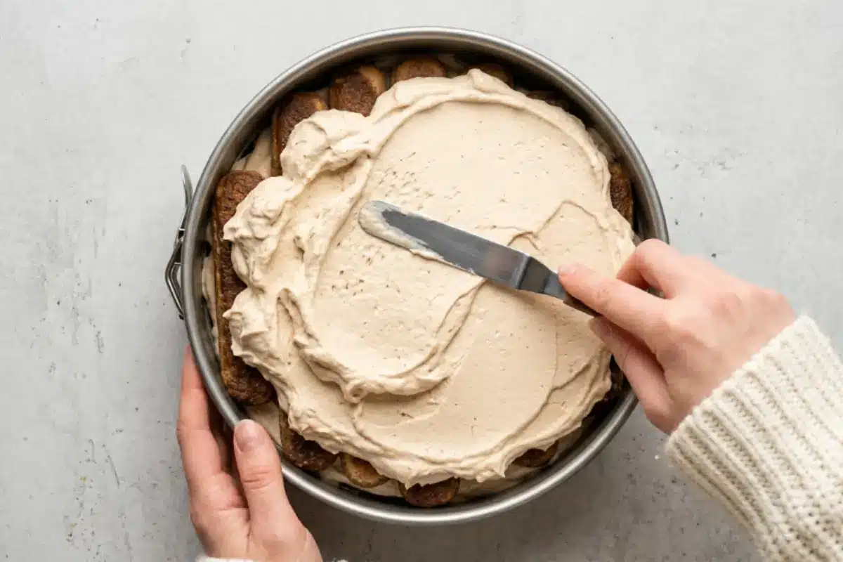 filling base For Nutella Tiramisu Cake A warm, top-down view of a hand gently using a metal spatula to spread a thick, velvety layer of espresso mascarpone cream over a base of coffee-soaked ladyfingers inside a springform pan, assembling the filling for a Nutella Tiramisu Cake.
