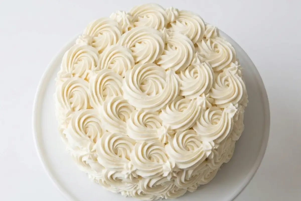 Frosting consistency guide 2 A beautiful round cake covered in thick, perfectly piped white buttercream rosettes on a simple white cake stand, illustrating the ideal texture for a frosting consistency guide.