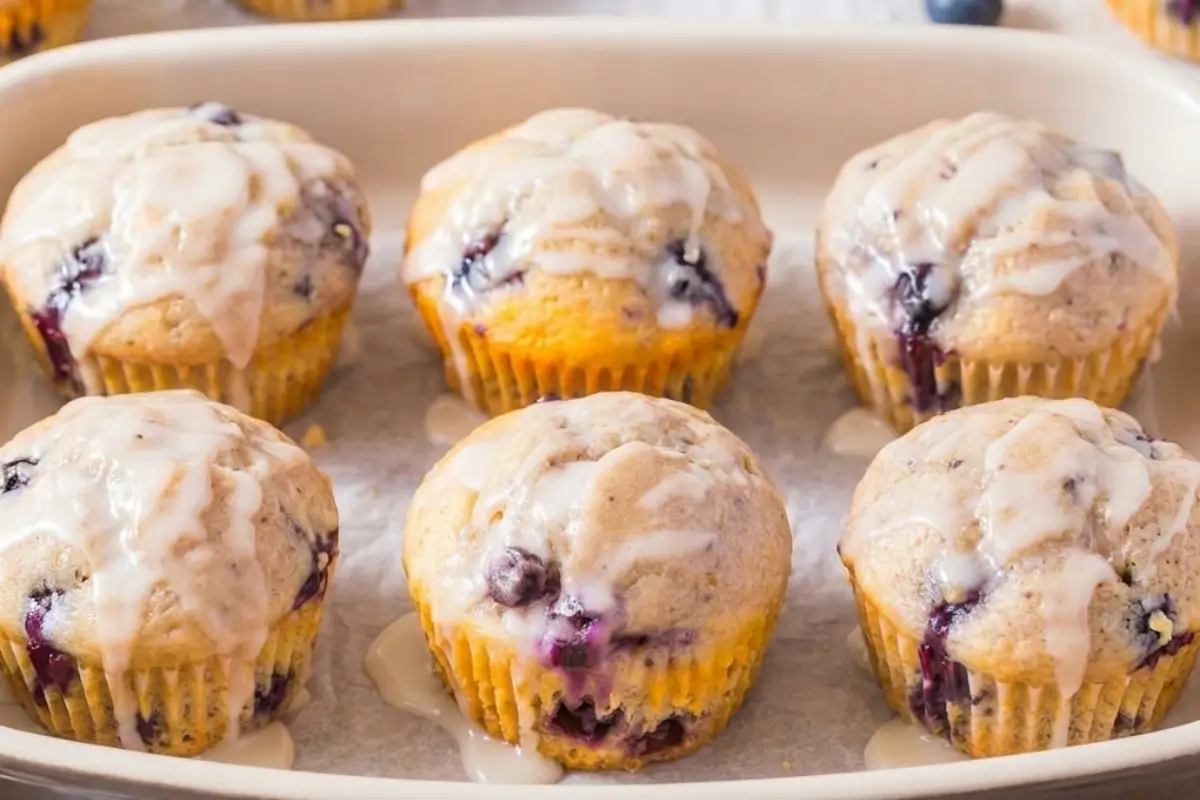 A freshly baked batch of six keto lemon blueberry cupcakes drizzled with a sweet glaze, resting in a rustic ceramic baking dish lined with parchment paper.