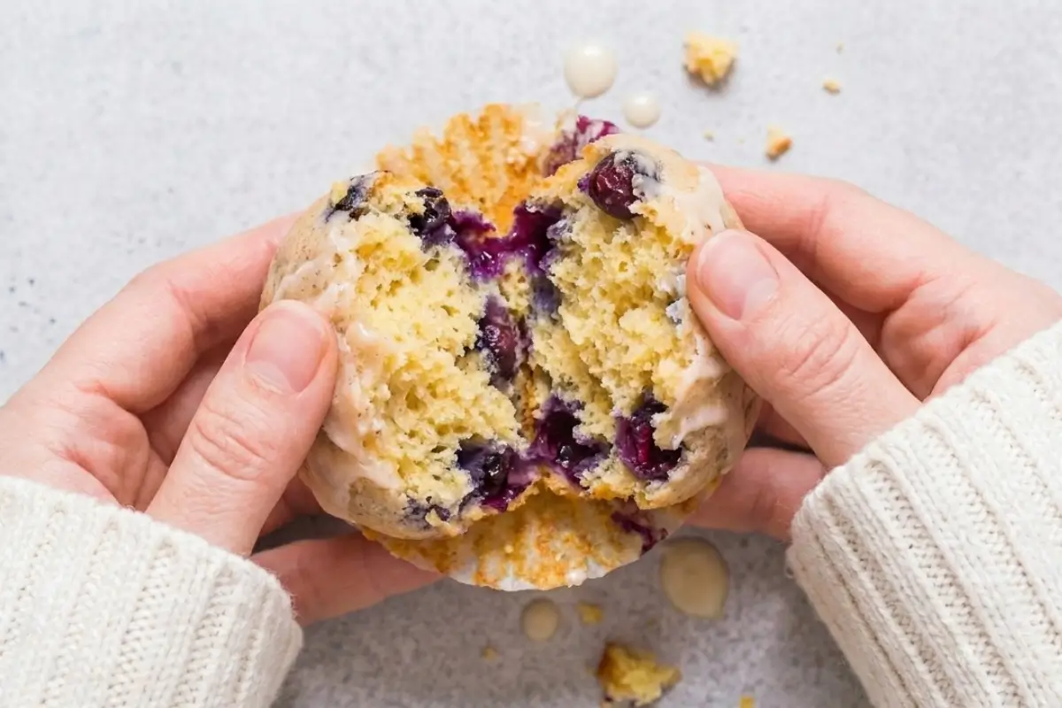 Lemon Blueberry Cupcakes recipe A pair of hands in a cozy knit sweater gently breaking apart a freshly baked, glazed treat from an easy lemon blueberry cupcakes recipe, revealing a tender crumb packed with juicy purple berries.