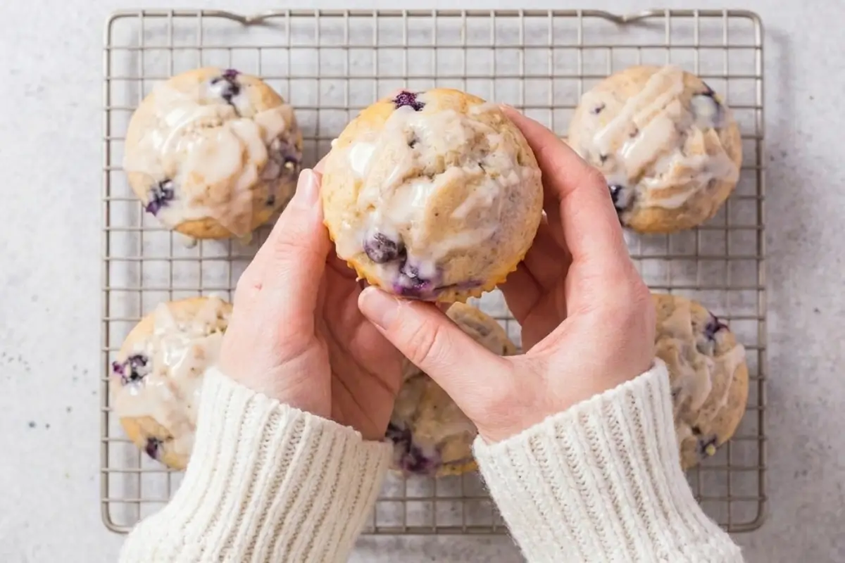 Perfect Keto Lemon Blueberry Cupcakes A pair of hands wearing a cozy cream knit sweater gently holding a freshly baked, glazed treat from a batch of perfect keto lemon blueberry cupcakes resting on a wire cooling rack.