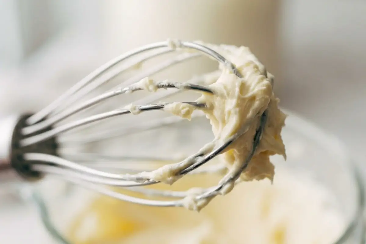 Which Frosting Should You Actually Use Lush, creamy frosting clinging to a metal whisk attachment above a glass mixing bowl, illustrating which frosting should you actually use for cakes.