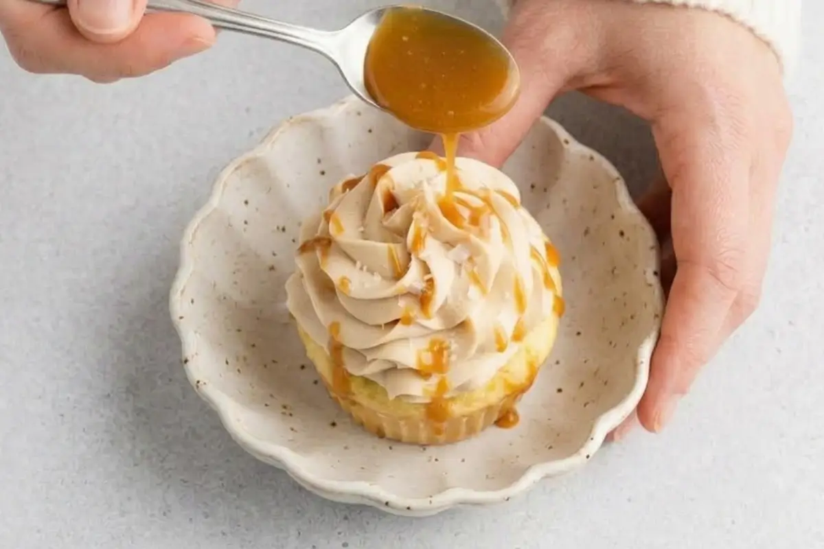 Why These Salted caramel filled cupcakes Work A pair of hands gently holds a warm, rustic ceramic bowl containing a beautifully frosted cupcake, while a silver spoon drizzles golden caramel sauce over the top to demonstrate exactly why these salted caramel filled cupcakes work so well