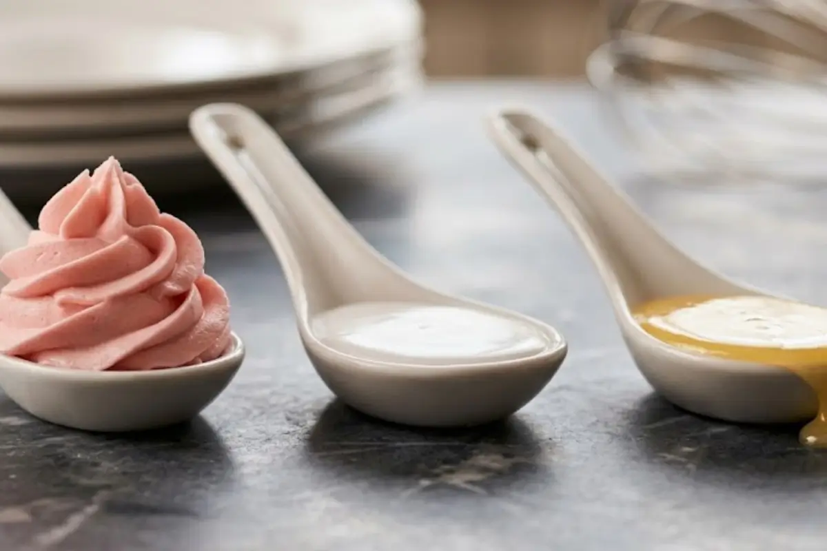 frosting vs icing vs glaze Three ceramic spoons comparing frosting vs icing vs glaze textures with a pink buttercream swirl, smooth white icing, and a drippy translucent glaze.